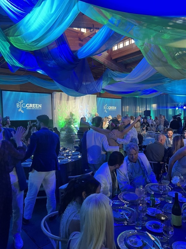 Guests dance between tables in a timber hall hung with blue and green fabric, with presentation screens at the front.