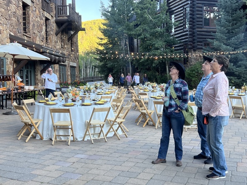Guests arrive at a stone-and-timber courtyard set with round tables, yellow napkins, flowers and strings of bistro lights.
