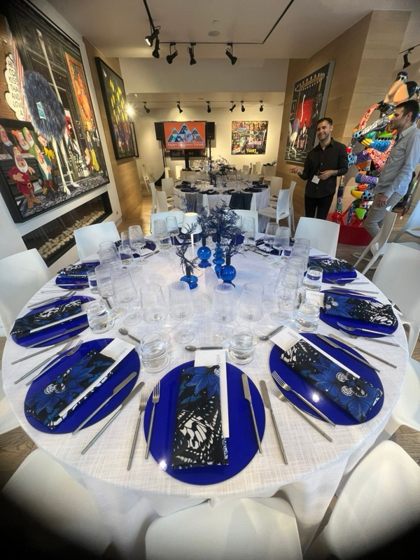A round dinner table in an art gallery, set with cobalt chargers, butterfly-print napkins, blue glass vases and printed menus, with more tables and a presentation screen beyond.