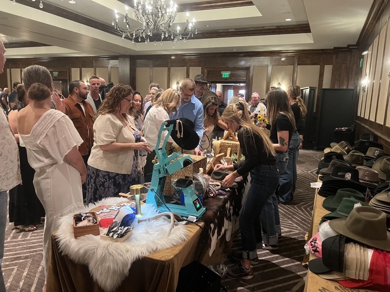 Guests gather at a hat-customising station in a hotel ballroom, with a heat press, trims and rows of felt hats.