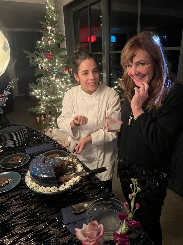 A chef cuts a glazed chocolate cake beside a smiling guest at a sequin-covered table, with a lit Christmas tree behind them.