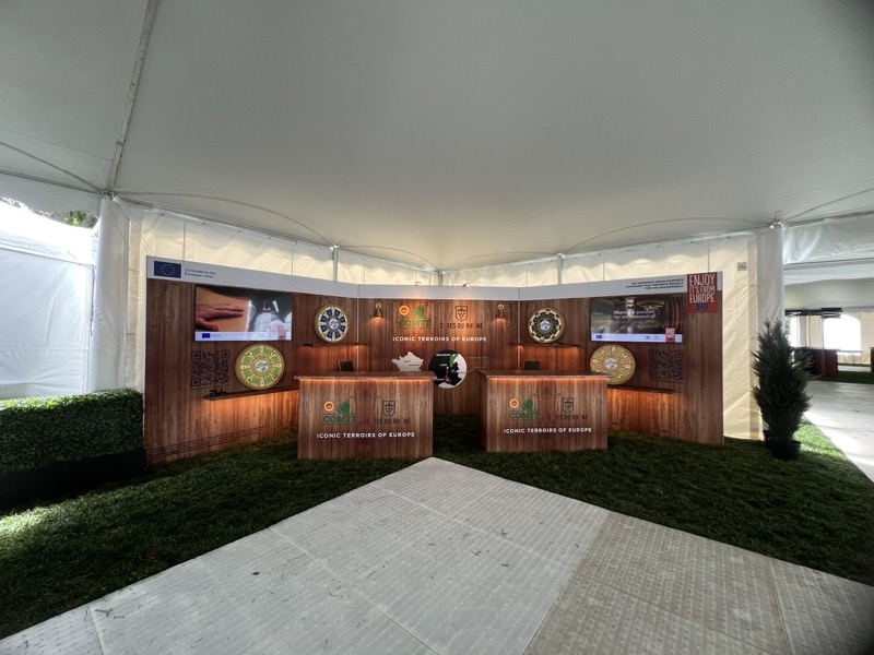 A wood-panelled tasting booth with two bars, screens and display shelves under a white tent, on a lawn floor.