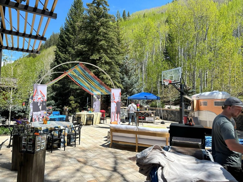 A sunny courtyard below aspen trees being set up for a party: an arch hung with rainbow ribbons, photo banners, colourful lounge tables, a basketball hoop and a vintage trailer bar.