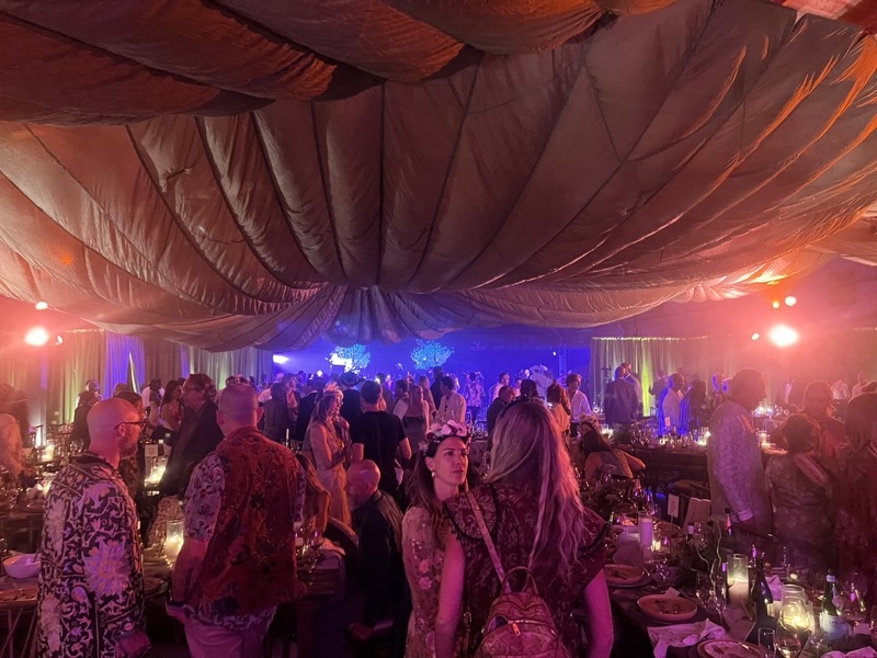 A crowded gala in a tent with billowing fabric overhead, warm lighting, candlelit tables and a lit stage beyond.