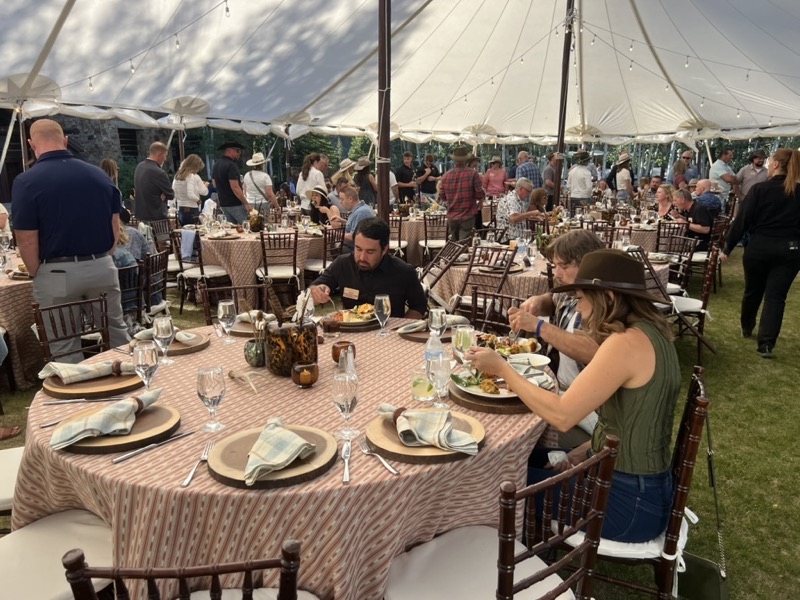 Guests in hats eat dinner at round tables with striped linens and wood chargers under a large white tent.