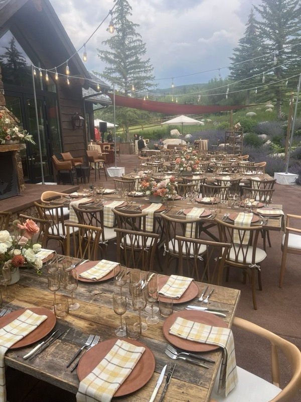 Long reclaimed-wood tables on a mountain-home terrace, set with terracotta chargers, plaid napkins and peach flowers under string lights.