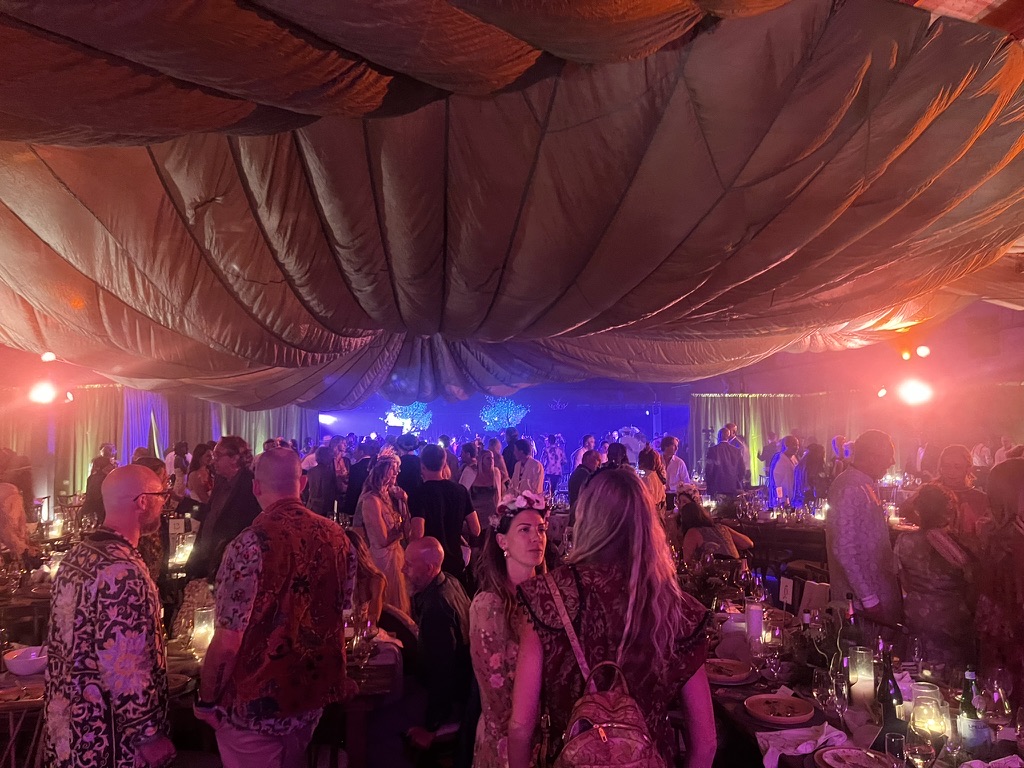 A crowded gala in a tent with billowing fabric overhead, warm lighting, candlelit tables and a lit stage beyond.