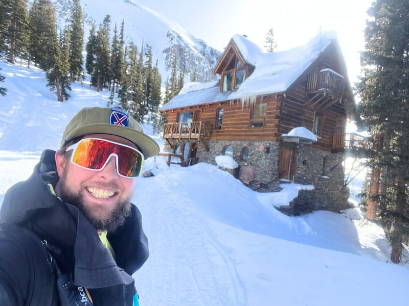 Max smiling on a snowy ski tour in mirrored goggles and a cap, a log cabin and steep peaks behind him.