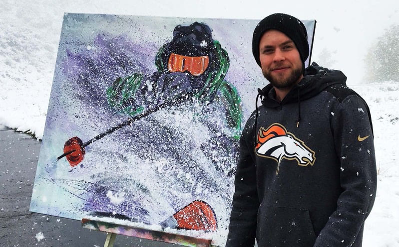 Max standing beside a large skier painting on an easel outdoors while snow falls.