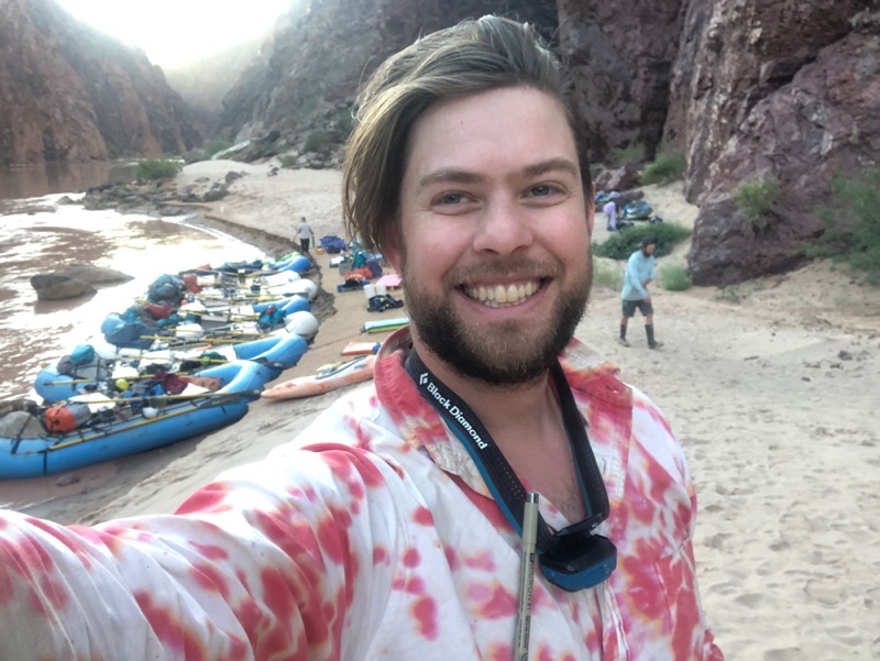 A selfie of Max at camp on a sandy beach, loaded rafts lined up along the river behind him.