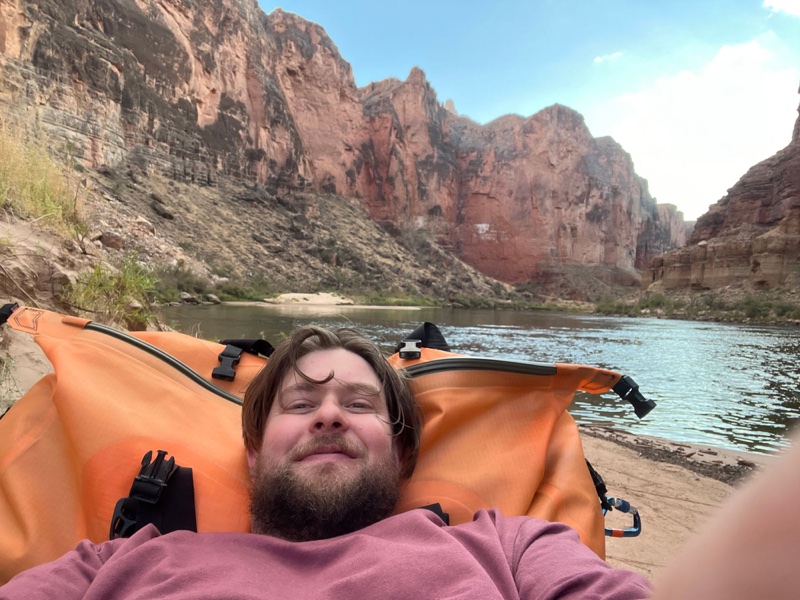A selfie of Max lying back against an orange dry bag on the riverbank, red canyon walls behind.