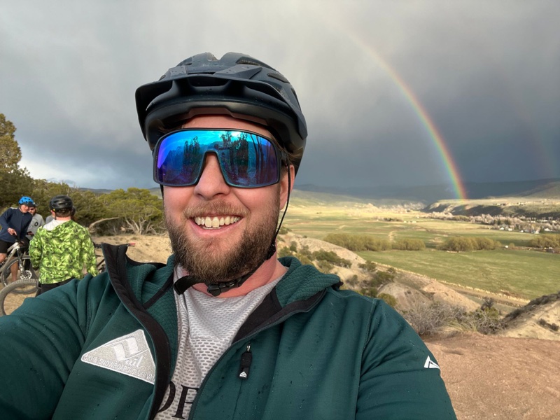 A selfie of Max in a bike helmet and blue mirrored sunglasses on a ridge above a green valley, a rainbow behind him and other riders nearby.
