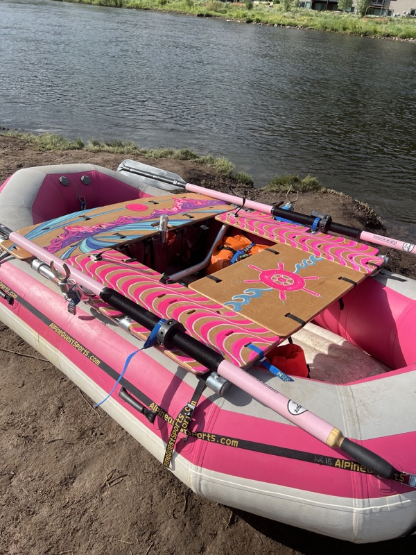 A pink and grey raft on a riverbank with a metal rowing frame, pink oars, and wooden deck boards painted with bright pink waves, a mountain scene and a ship's wheel.
