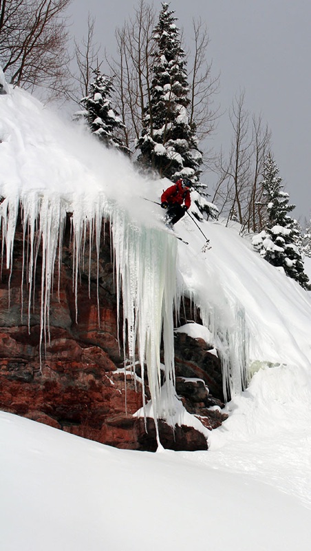 Max in a red jacket skiing off a cliff band hung with long icicles, into deep powder below snowy trees.