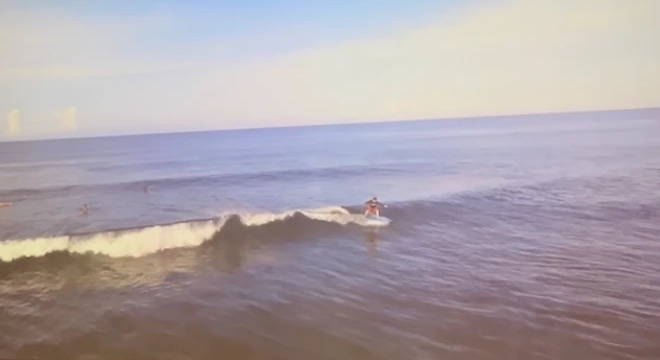 Max riding a small wave on a surfboard, seen from above, with open ocean behind.