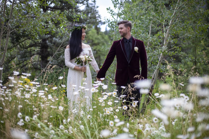 Max and Charlotte holding hands in a field of white daisies among aspens; Charlotte in a white lace dress with a flower crown and bouquet, Max in a burgundy velvet jacket.